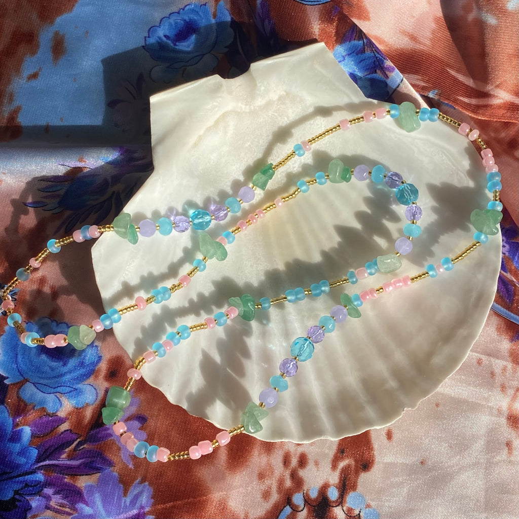 A gold, blue, and pink waist bead with green aventurine gemstone and lilac and blue faceted beads, displayed on a white shell.