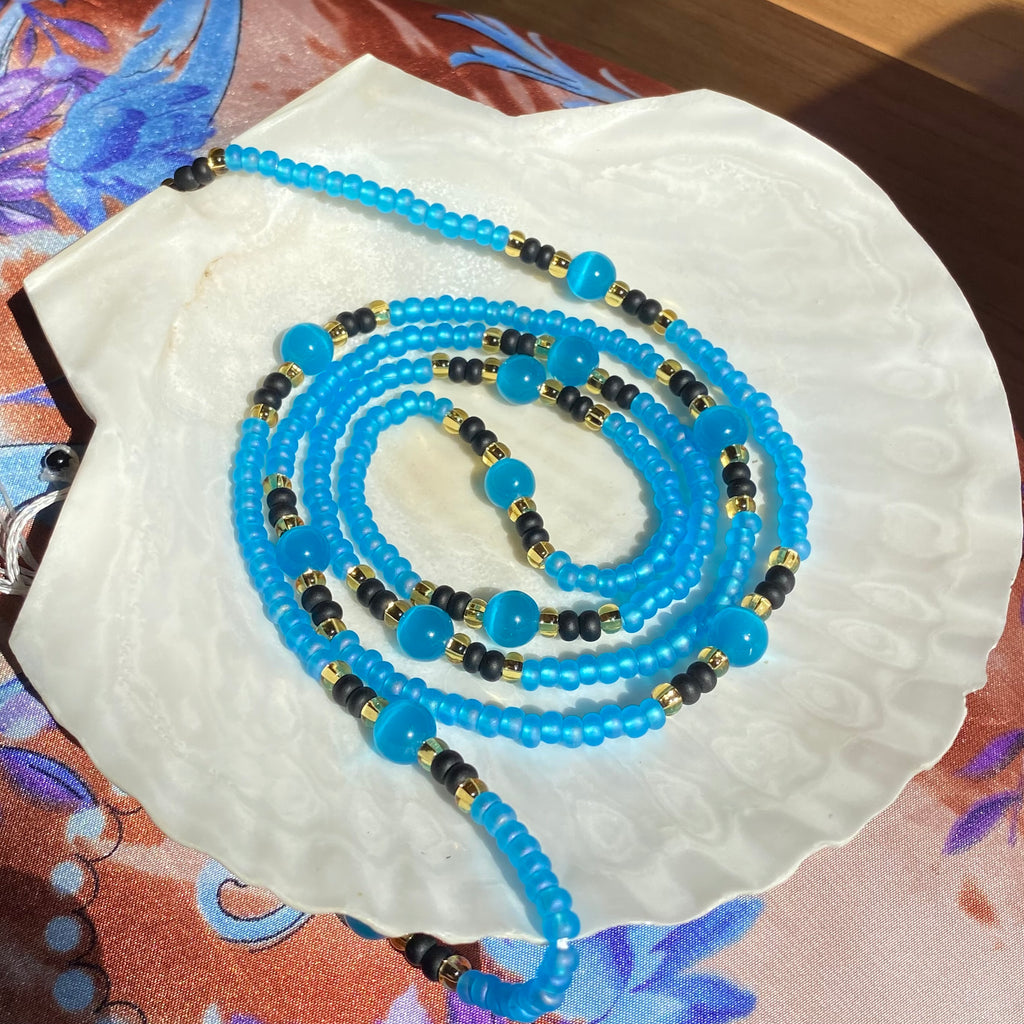 blue black a and gold waist beads with blue cat's eye beads displayed on a white shell.