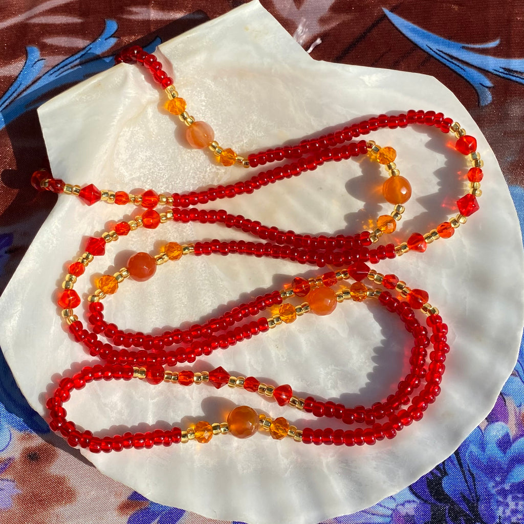 A red waist bead with facetted carnelian and orange beads displayed on a white shell