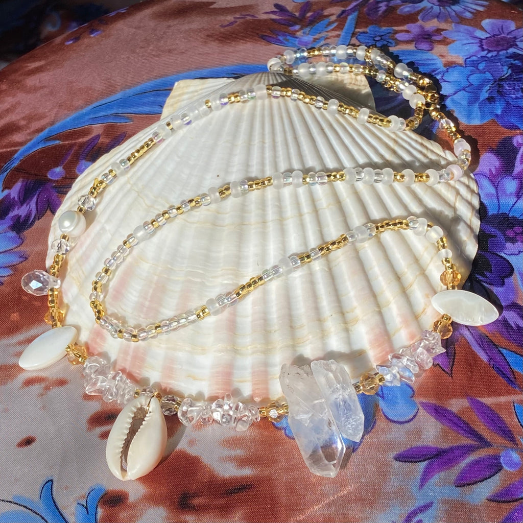 clear and gold waist beads with clear quartz, shells and pearls displayed on a white shell and a floral background