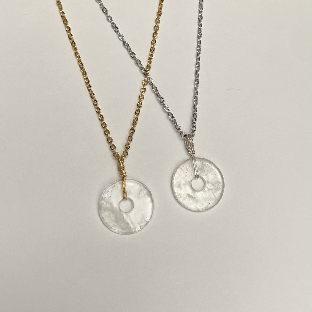 Two necklaces laid out on a flat surface, one with a gold-toned chain and pendant, and the other with a silver-toned chain and pendant. The pendants are clear quartz discs.