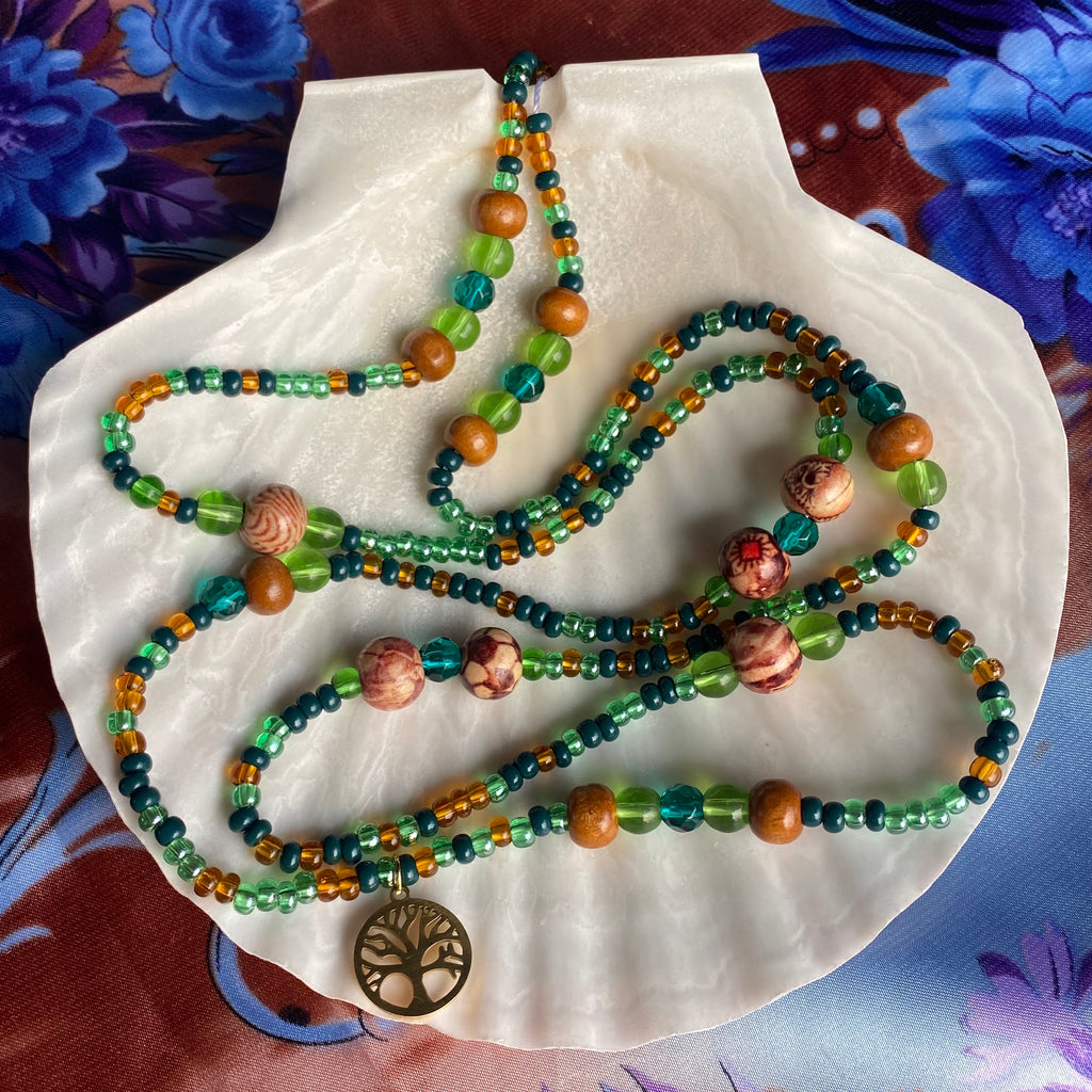 A green and brown waist bead with wood beads and a gold colored tree of life pendant displayed on a white shell