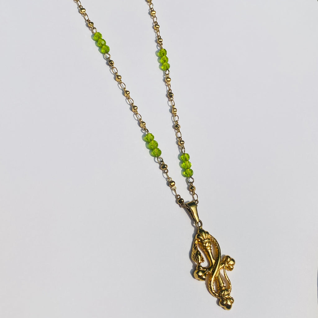 Gold necklace with green beads and a snake on a cross pendant displayed on a white background.
