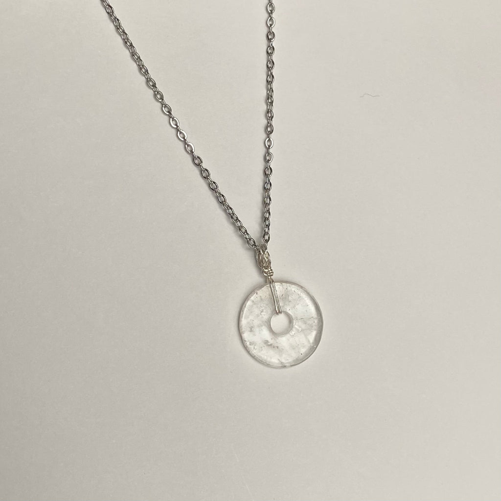 a necklace layed out on a flat surface with a silver-toned chain and a clear quartz disc pendant.