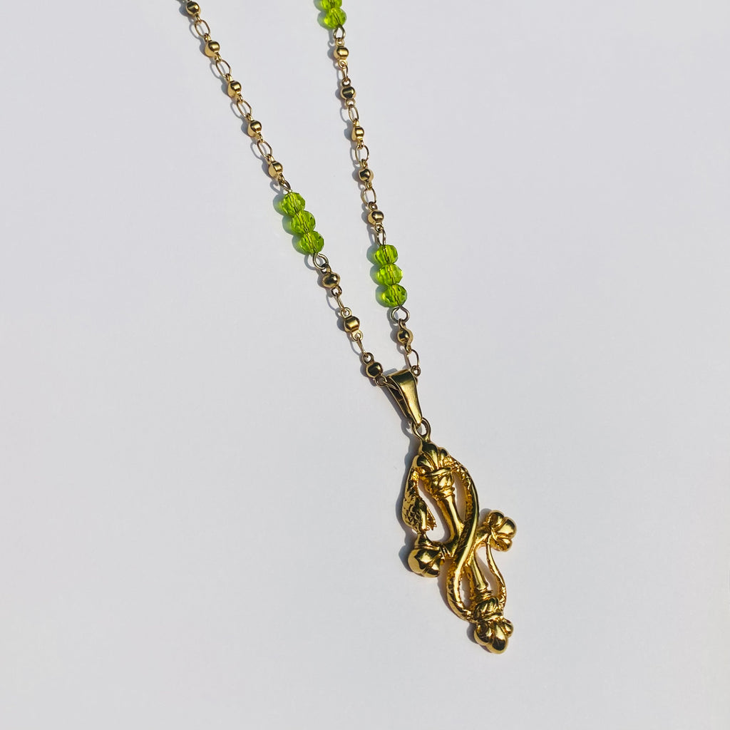 Gold necklace with green beads and a snake on a cross pendant displayed on a white background.