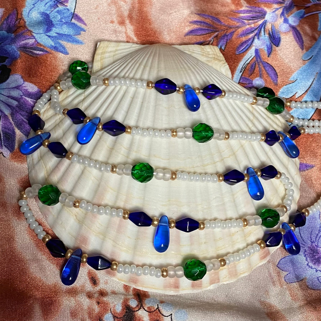 A pearly white and gold waist bead with royal blue drops and deep blue prisms and green facetted beads displayed on a seashell.